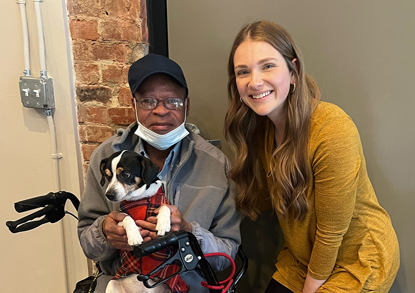 woman posing with a man in his wheelchair holding up his small dog